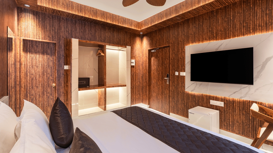 Modern hotel bedroom with textured wooden walls, white bedding, a dark grey bed runner, and wall-mounted television.