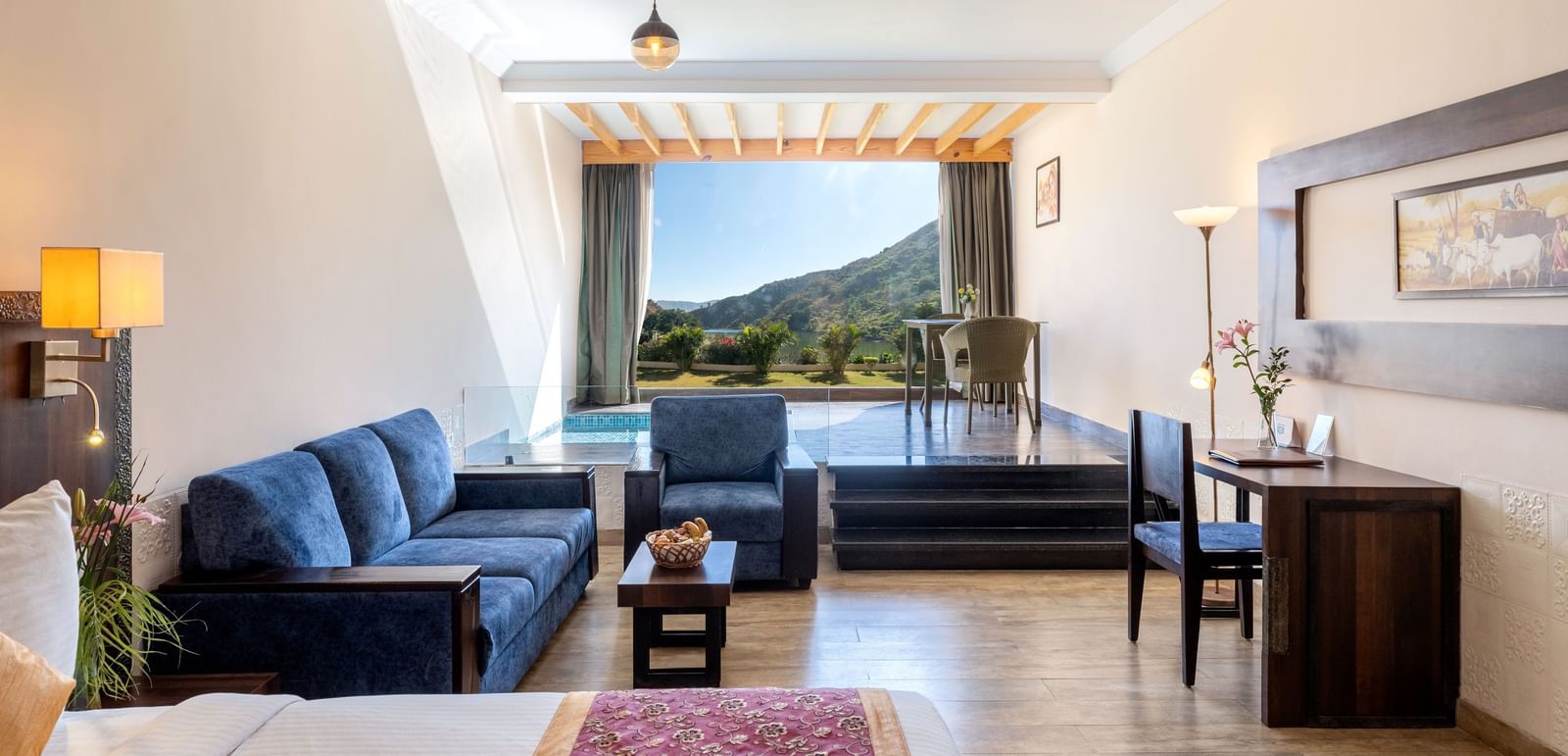 A living area with a blue couch, a neatly arranged bed, and a window with mountain view - Hotel Udai Median, Udaipur