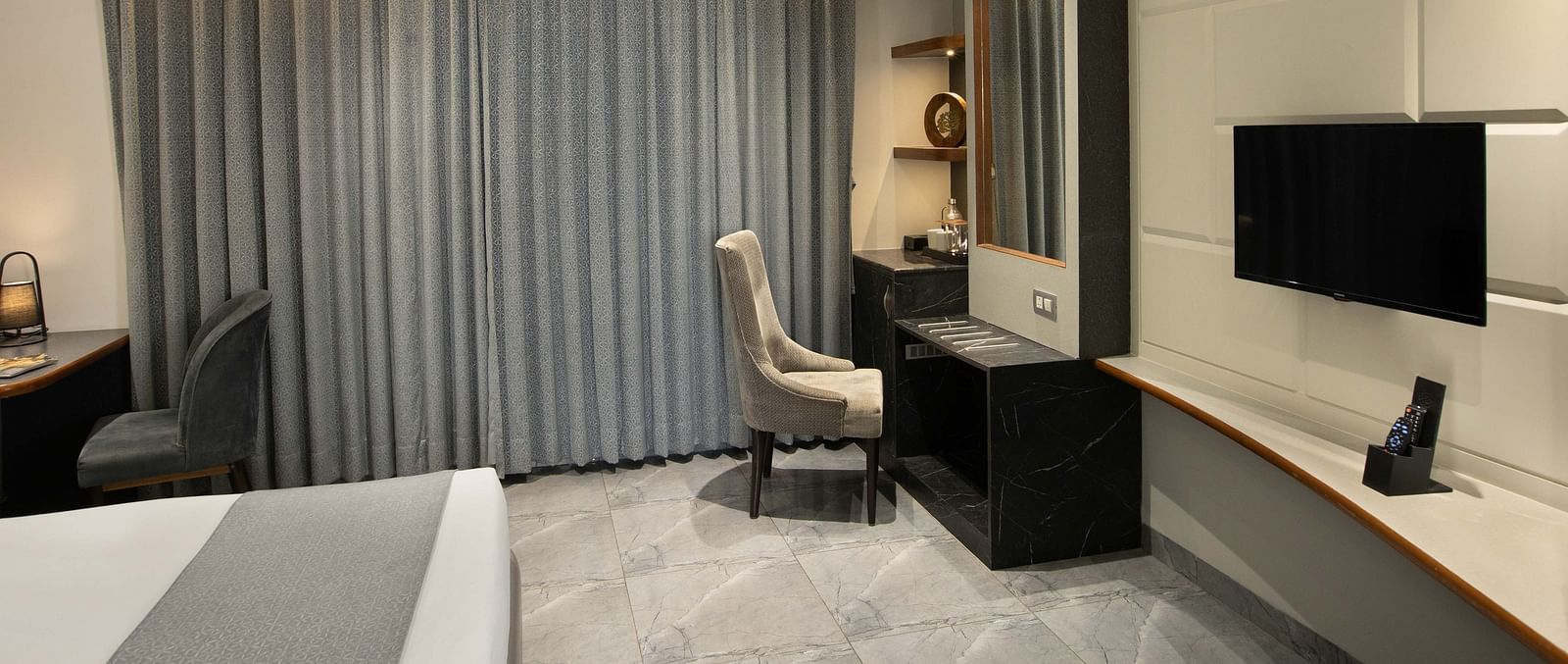 Image of the Coral Deluxe Room at Coral Reef Hotel & Spa in Port Blair featuring chair in front of the mirror next to the window and a TV mounted on the wall in front of the bed