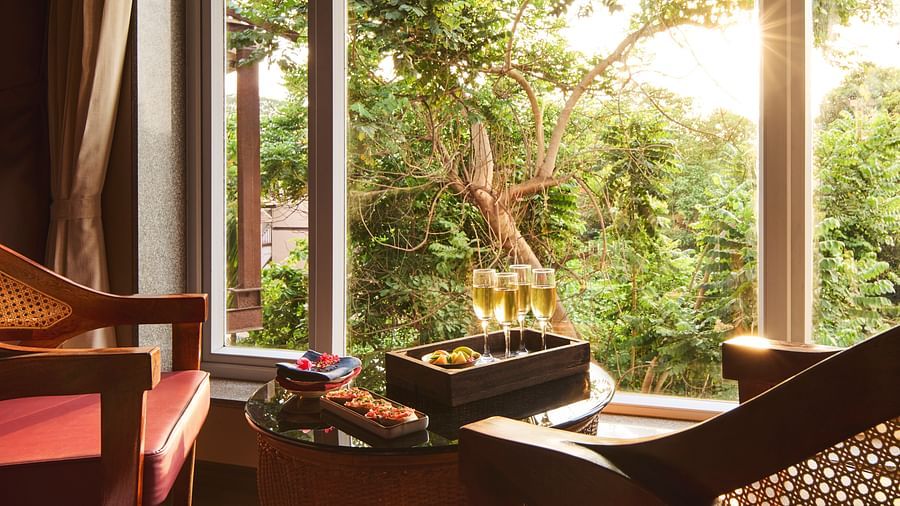 A table with four champagne flutes and snacks sits in the living room of Super Deluxe Family Room by a large window, offering a sunny view of lush green trees at DGV Resort by Treat, Silvassa.