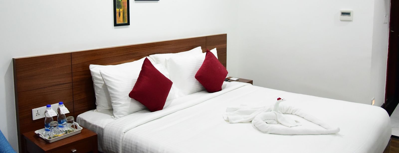 The luxurious comfort club room at The Royal Comfort hotel, featuring a plush bed with white linens and two deep red accent pillows, elegantly decorated walls, and a bedside table with amenities.