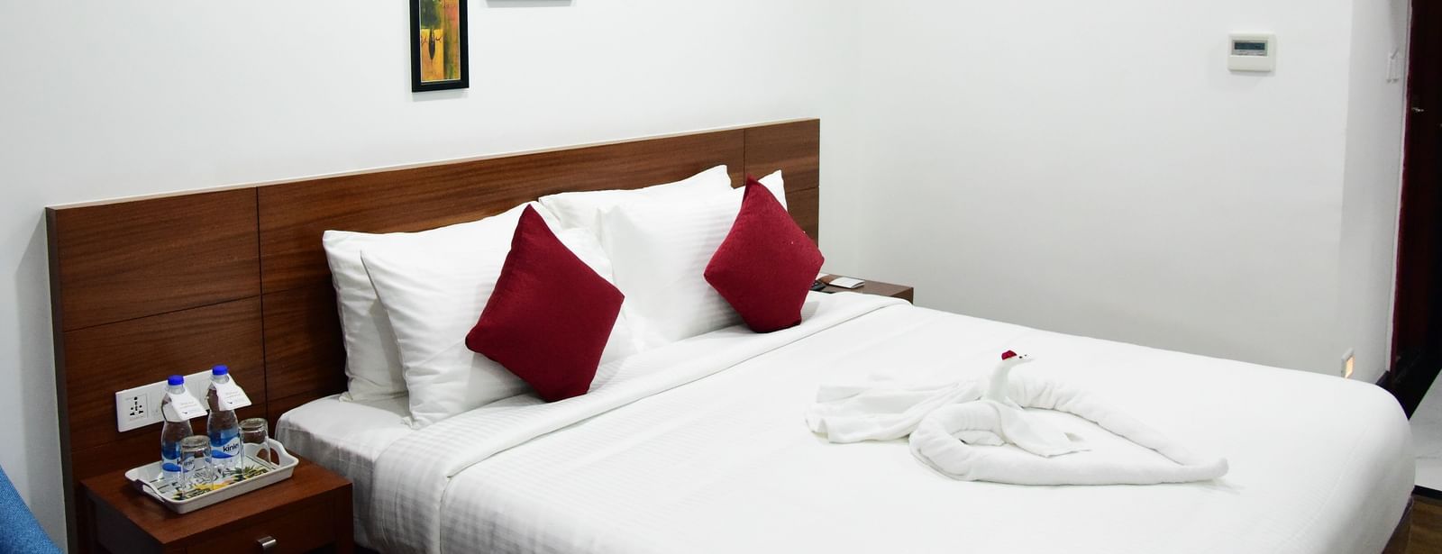 The luxurious comfort club room at The Royal Comfort hotel, featuring a plush bed with white linens and two deep red accent pillows, elegantly decorated walls, and a bedside table with amenities.