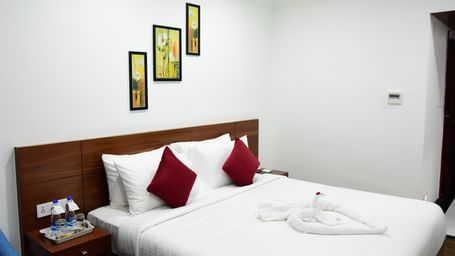 The luxurious comfort club room at The Royal Comfort hotel, featuring a plush bed with white linens and two deep red accent pillows, elegantly decorated walls, and a bedside table with amenities.