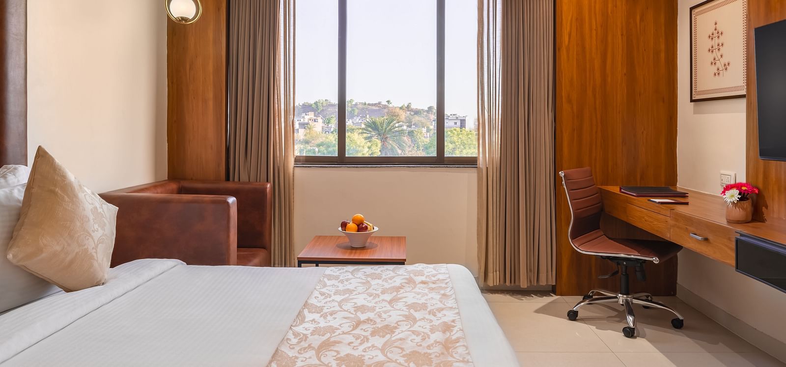 A well-lit hotel room features a comfortable bed, a desk with a chair, and a window with curtains offering a cityscape view - Lords Eco Inn Ambaji.