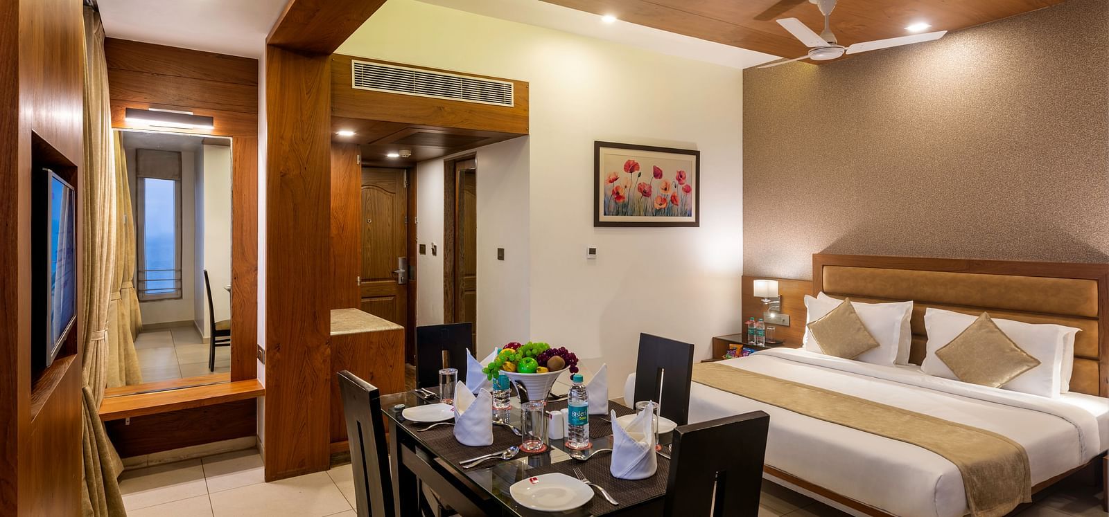 A comfortable and modern hotel suite featuring a king-sized bed with neutral-toned linens, a dark wood dining table set for four with colorful flowers, a wall-mounted television, and a glimpse into an adjoining area with a mirror and closet space.