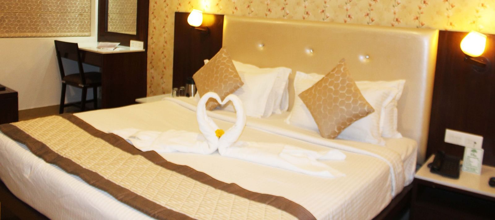 A warmly lit hotel room with a queen size bed, white bedding and a towel art swan on the bedspread - TGI Apple Inn, Jaipur 