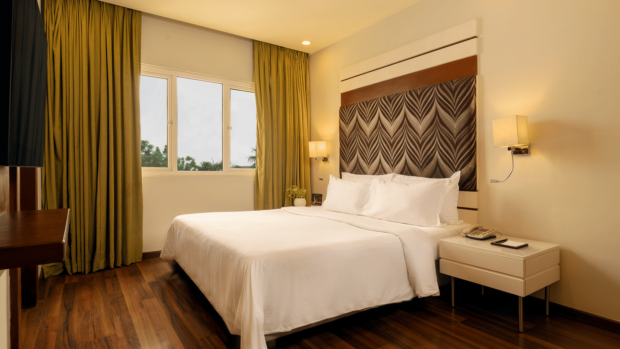 A room with a queen bed, side table and curtained windows overlooking the greenery at The Residency Karur.