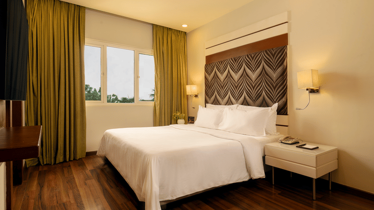 A room with a queen bed, side table and curtained windows overlooking the greenery at The Residency Karur.
