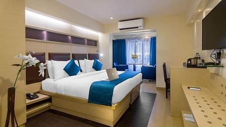 Side view of a spacious room with large bed, pillows, sheets, window with curtains, AC above, and wall-mounted TV at Oriental Aster, Mumbai.