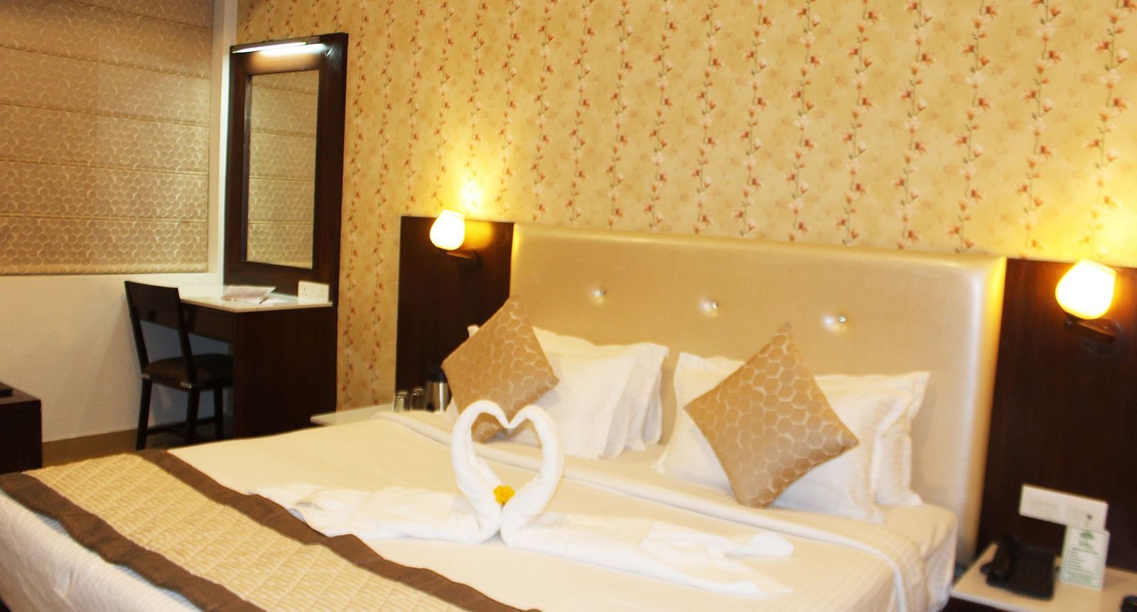 A warmly lit hotel room with a queen size bed, white bedding and a towel art swan on the bedspread - TGI Apple Inn, Jaipur 