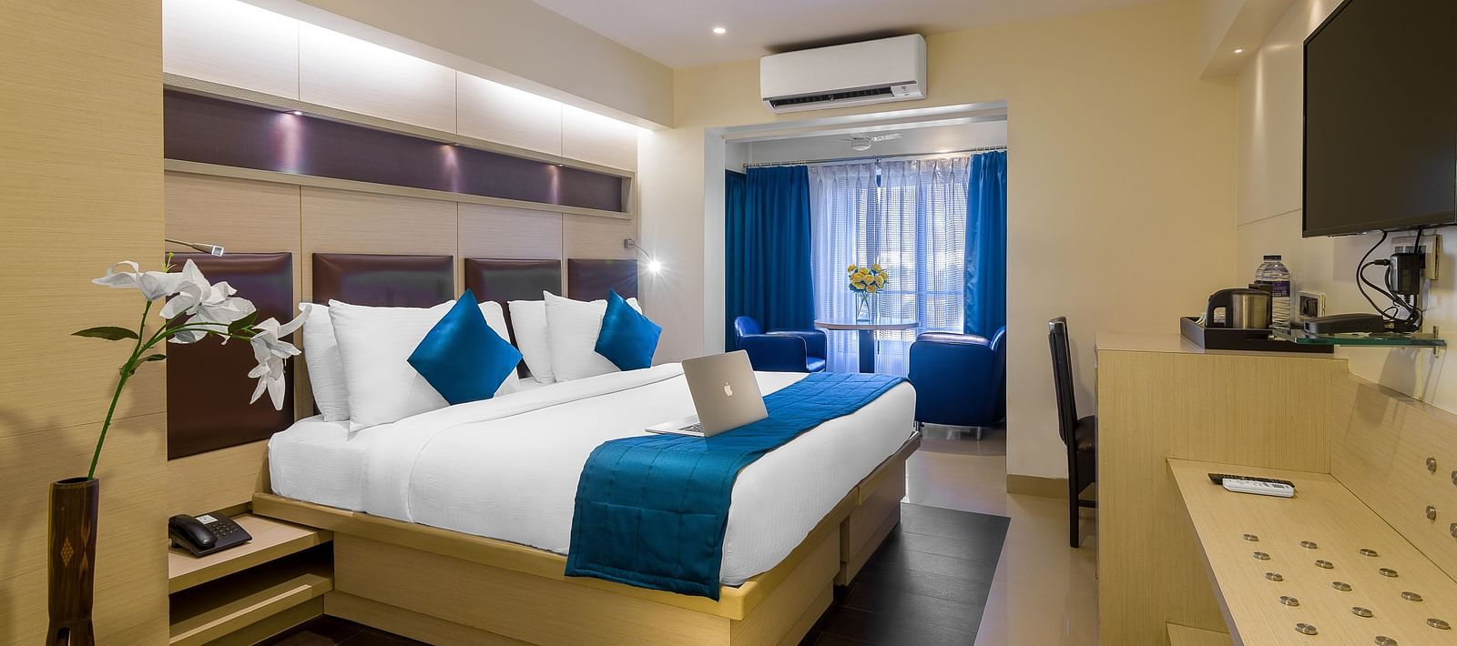 Side view of a spacious room with large bed, pillows, sheets, window with curtains, AC above, and wall-mounted TV at Oriental Aster, Mumbai.