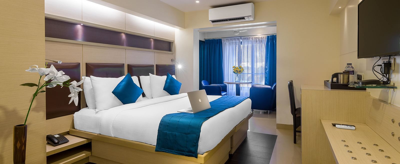 Side view of a spacious room with large bed, pillows, sheets, window with curtains, AC above, and wall-mounted TV at Oriental Aster, Mumbai.