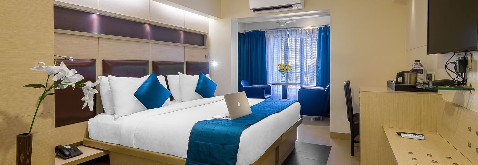 Side view of a spacious room with large bed, pillows, sheets, window with curtains, AC above, and wall-mounted TV at Oriental Aster, Mumbai.