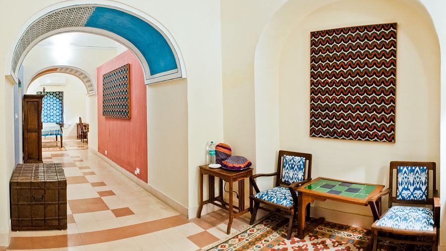 Hotel hallway with arched openings, a seating area with chairs and a table on a rug, artwork, and a chest at Tijara Fort-Palace, Alwar.