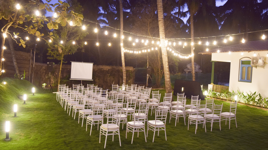 Outdoor night event setting with white chairs, a projector screen, and decorative string lights over a lawn at Ibex Resorts, Coimbatore (Kakarla).