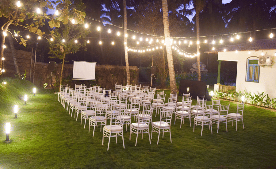 Outdoor night event setting with white chairs, a projector screen, and decorative string lights over a green lawn.