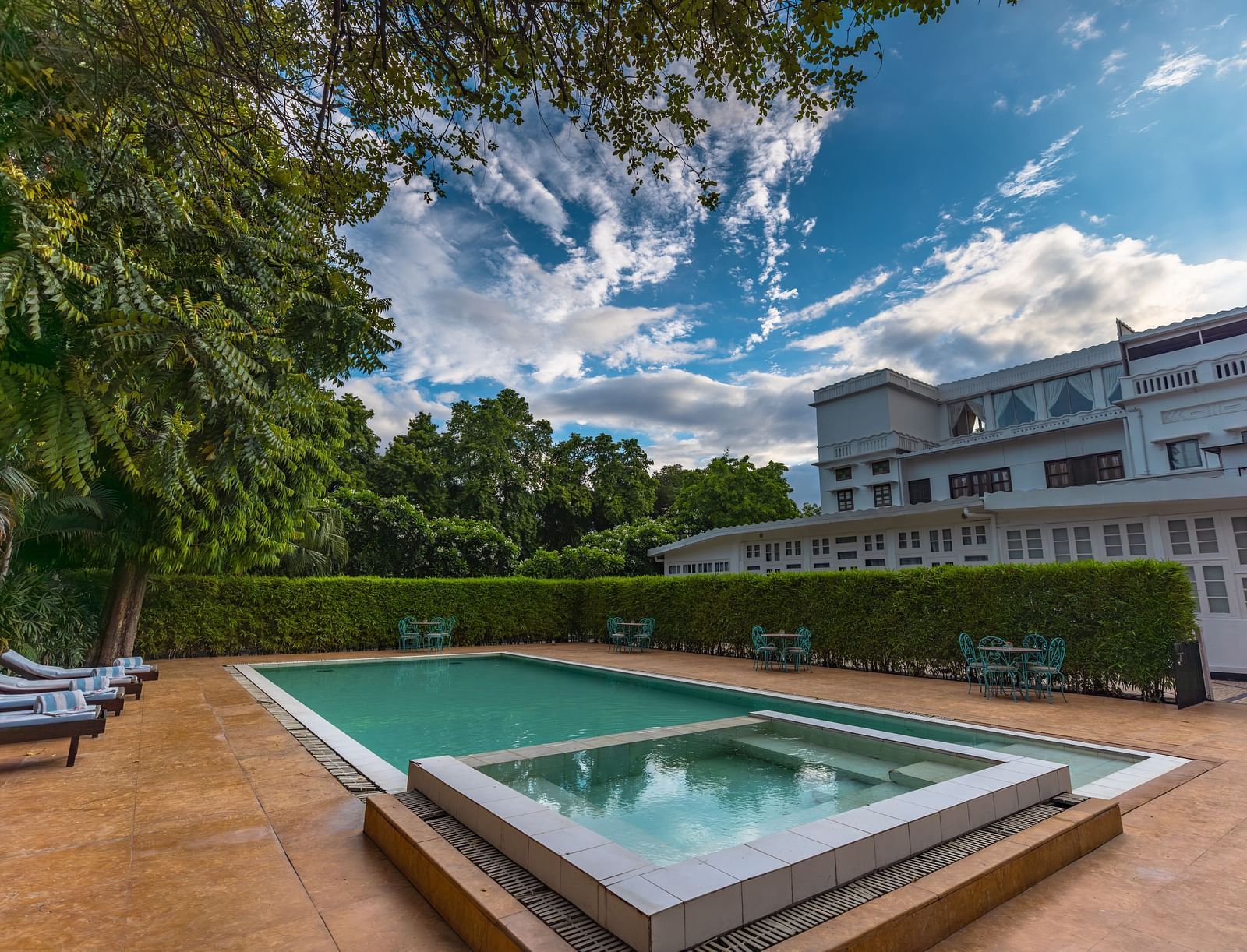 Outdoor swimming pool area of Saraca Hotel, Lucknow with lounging chairs placed around it and the hotel building in the backdrop on a cloudy day