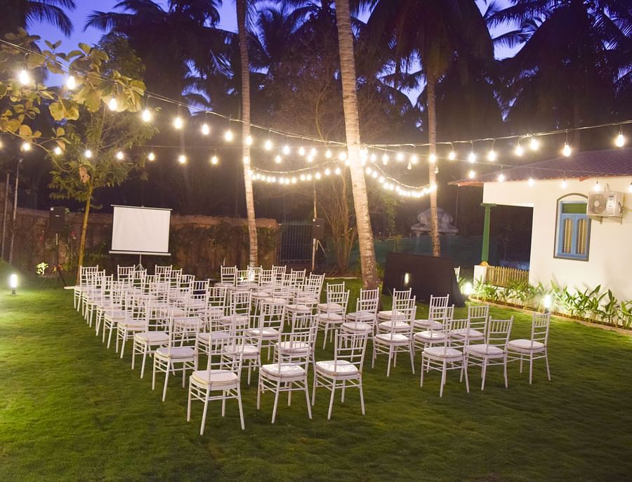 Outdoor night event setting with white chairs, a projector screen, and decorative string lights over a green lawn.