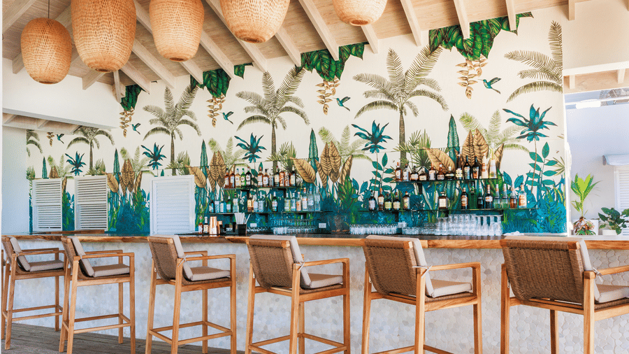 A outdoor bar area with wooden chairs, counter, tropical plant murals, and shelves of different types and brands of alcohol at Tamarind Hills Resort and Villas, Antigua.