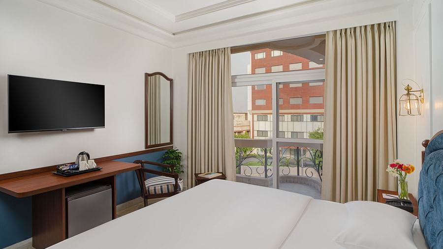 A spacious bed with a wall-mounted television above a wooden table, alongside a large window with the curtains drawn to the side | Hotel Ganga Maiya