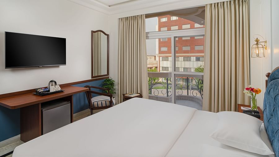 A spacious bed with a wall-mounted television above a wooden table, alongside a large window with the curtains drawn to the side | Hotel Ganga Maiya