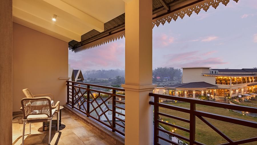 A balcony view at dusk or dawn, showcasing two wicker chairs and a small table on a tiled floor, with a decorative brown railing overlooking a green lawn and buildings with warm lights in the distance under a soft, colourful sky - Voyage The Solitaire Blis