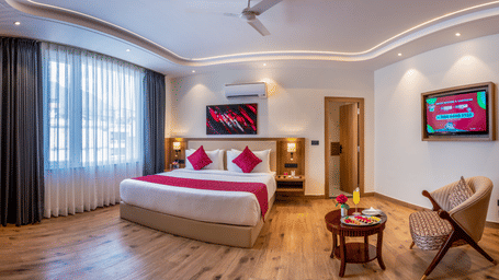 A bright room at Lords Inn Katra containing a king size bed, a large window, an armchair with a side table, and a wall television.