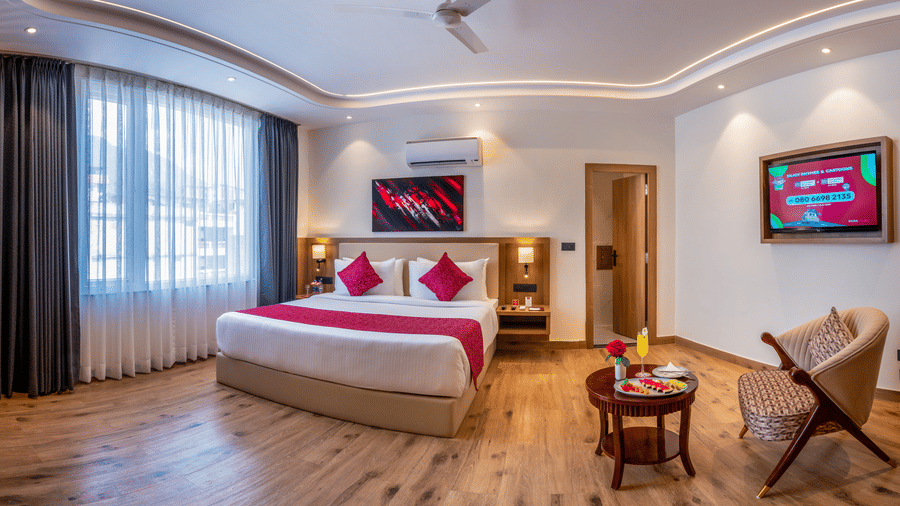 A bright room at Lords Inn Katra containing a king size bed, a large window, an armchair with a side table, and a wall television.