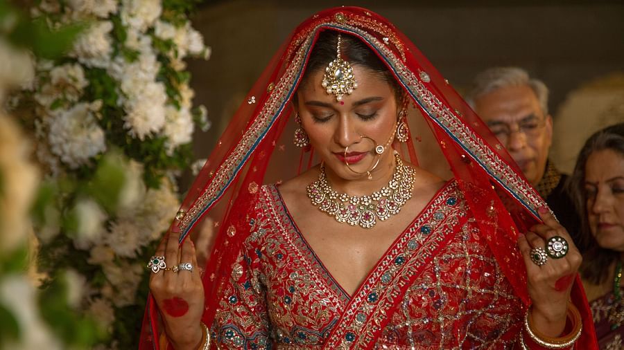 A bride in traditional indian attire, featuring a red lehenga and veil, adorned with intricate gold jewelry, looks down modestly during a wedding ceremony.