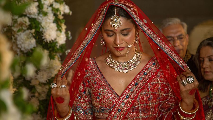 A bride in traditional indian attire, featuring a red lehenga and veil, adorned with intricate gold jewelry, looks down modestly during a wedding ceremony.
