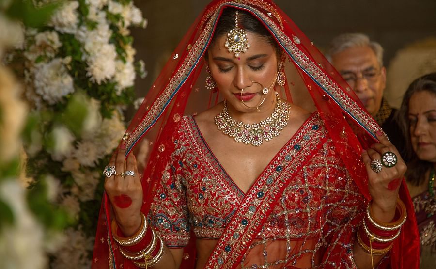 A bride in traditional indian attire, featuring a red lehenga and veil, adorned with intricate gold jewelry, looks down modestly during a wedding ceremony.