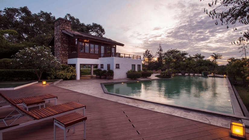 A modern luxury villa with a stone and white exterior beside an inviting swimming pool, surrounded by lush greenery and deck chairs at sunset.
