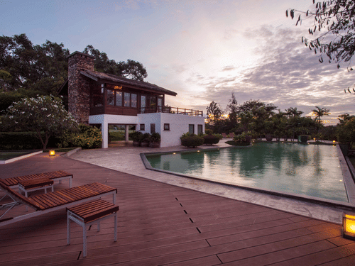 A modern luxury villa with a stone and white exterior beside an inviting swimming pool, surrounded by lush greenery and deck chairs at sunset.