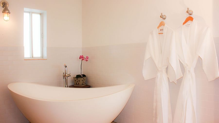 a bathroom at casa morada with a bathtub, hanging bathrobes , a flower vase and a small window.