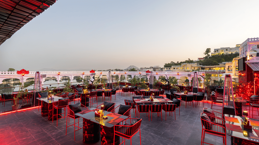 Wide exterior shot of a rooftop bar at dusk, with numerous red-lit tables and distant buildings on a hillside.