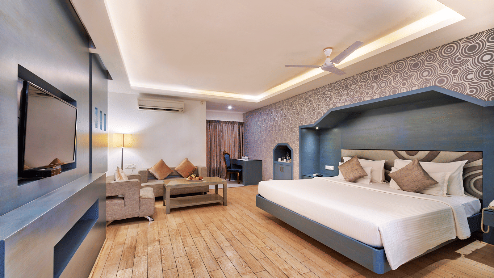 A modern hotel bedroom with a large bed, a sofa, a coffee table, and a wall-mounted television - Grande Bay Resort & Spa, Mamallapuram