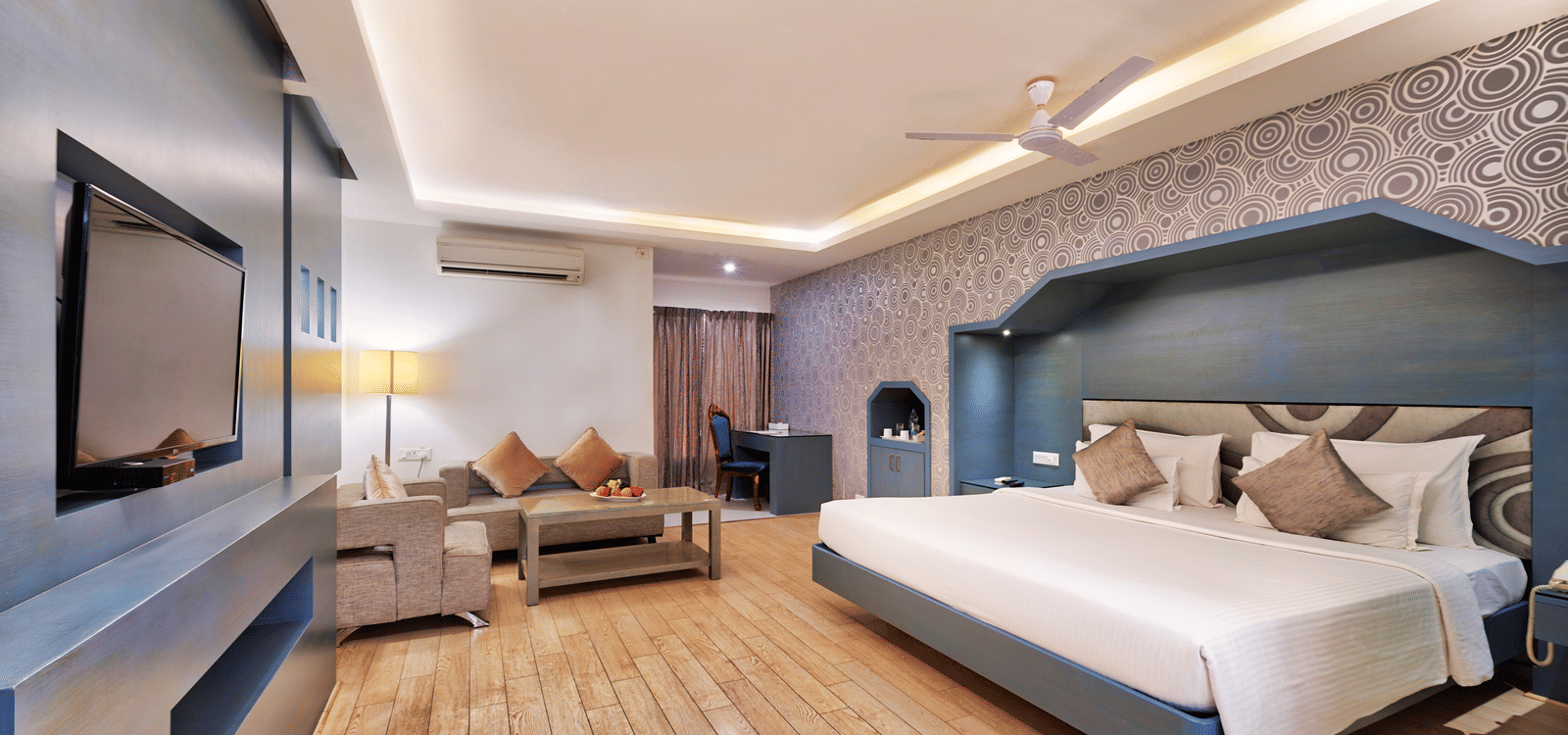 A modern hotel bedroom with a large bed, a sofa, a coffee table, and a wall-mounted television - Grande Bay Resort & Spa, Mamallapuram