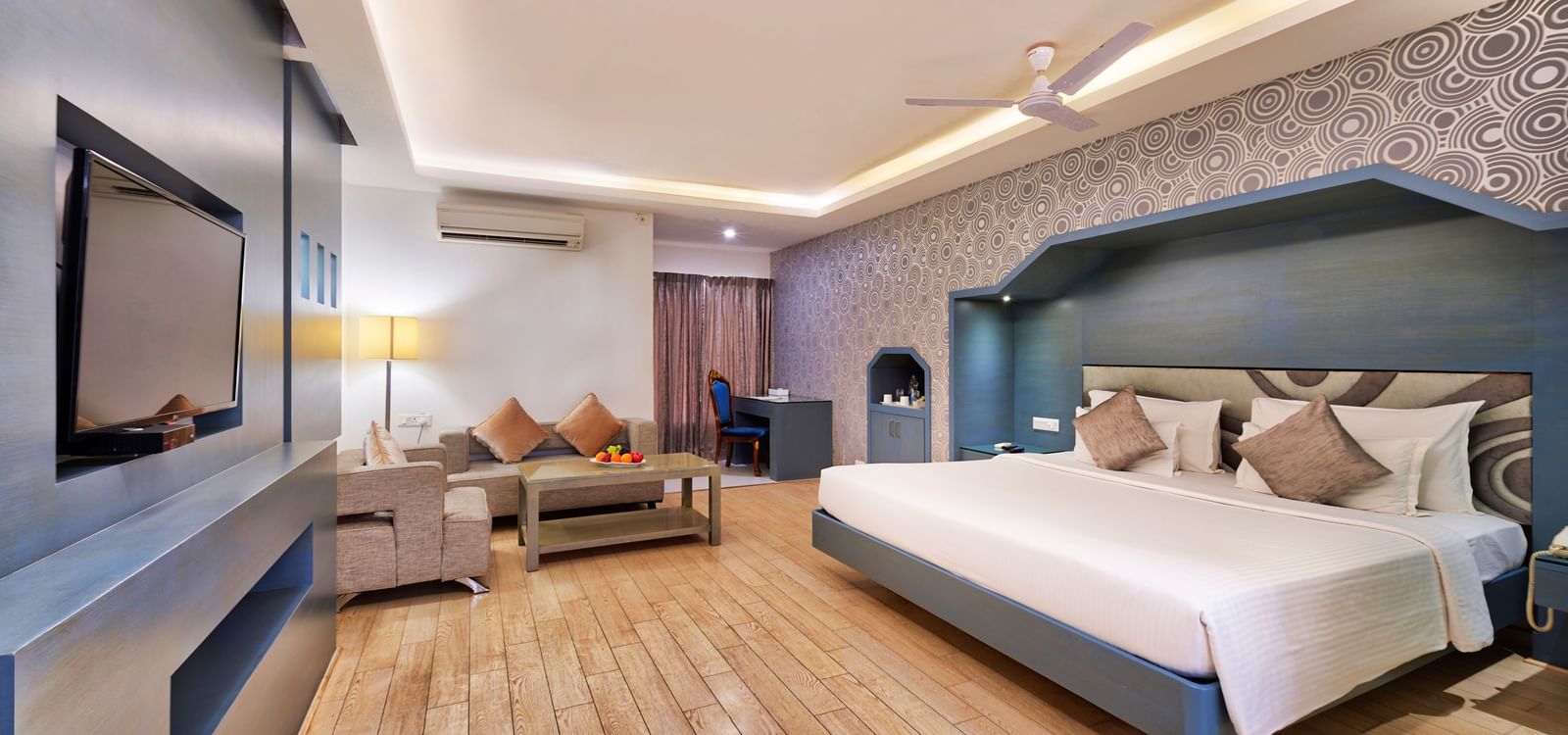 A wide-angle view of a hotel room with a large bed, a television cabinet, and a seating area | Grande Chalet | Grande Bay Resort & Spa, Mamallapuram