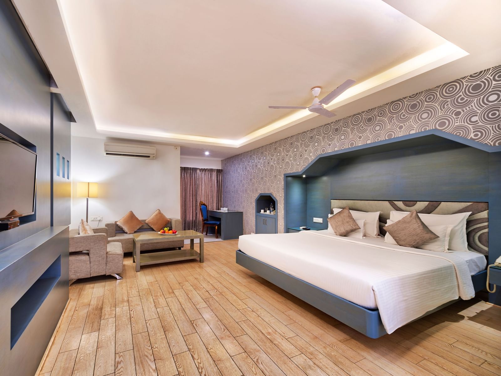A wide-angle view of a hotel room with a large bed, a television cabinet, and a seating area | Grande Chalet | Grande Bay Resort & Spa, Mamallapuram