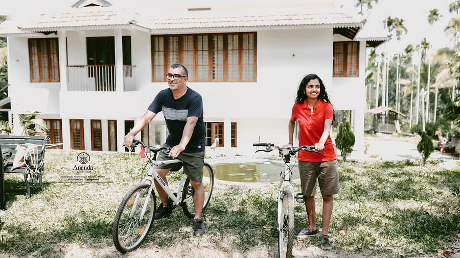 A man and a woman sitting on bicycles in front of the facade of Ananda Holiday Home, Wayanad