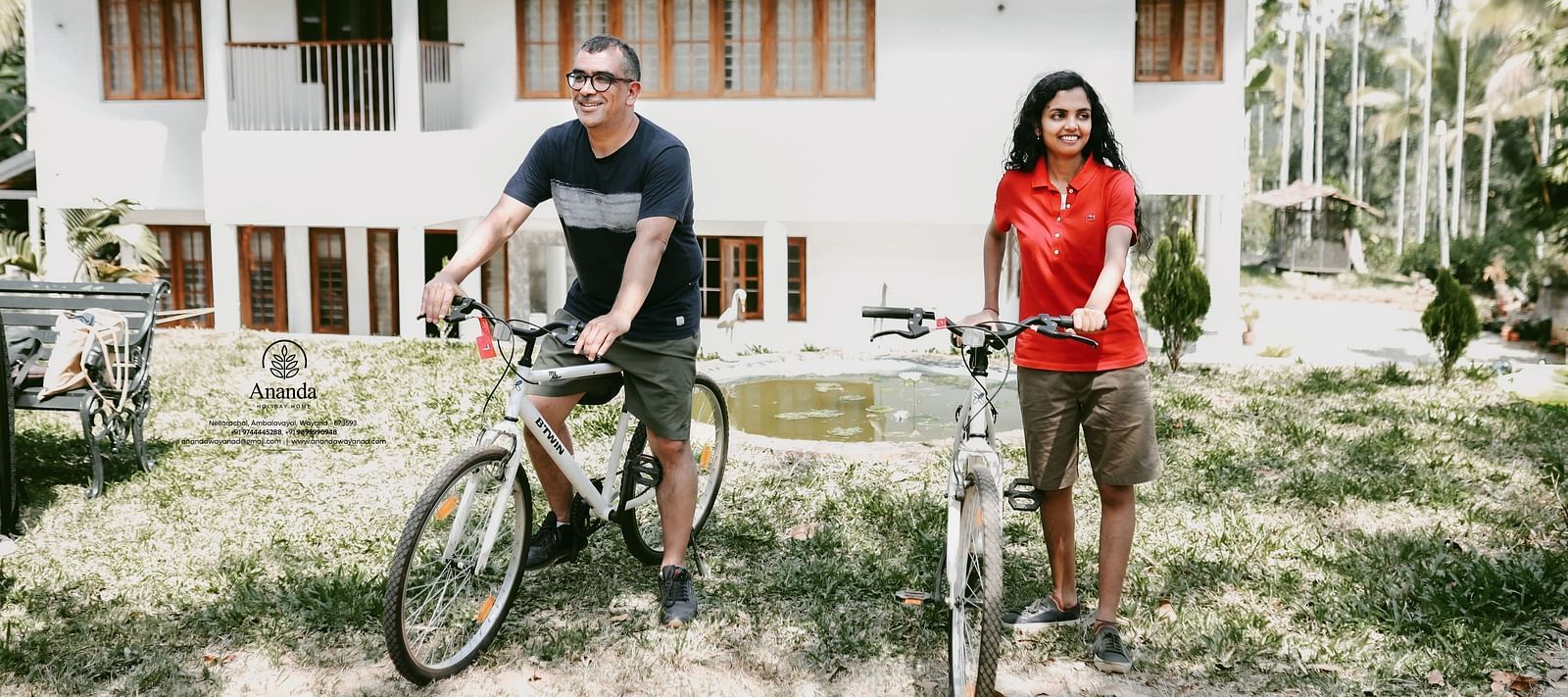 A man and a woman sitting on bicycles in front of the facade of Ananda Holiday Home, Wayanad
