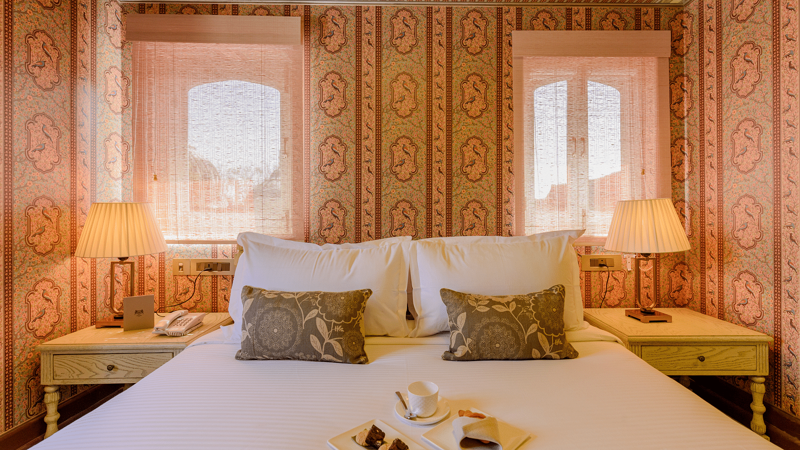 Deluxe rooms in Surya Haveli Jaipur featuring a plush bed with cookies and bed tea on the sheet and well decorated walls along with a couple of lamps on both sides of the bed on the nightstands
