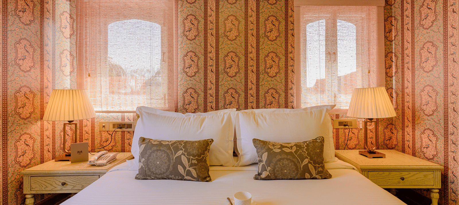 Deluxe rooms in Surya Haveli Jaipur featuring a plush bed with cookies and bed tea on the sheet and well decorated walls along with a couple of lamps on both sides of the bed on the nightstands