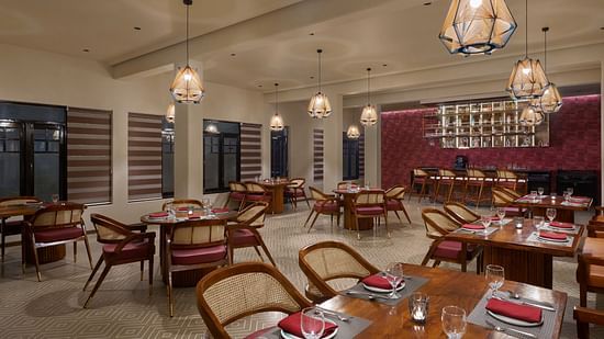 Elegant indoor restaurant with warm lighting, wooden furniture, red cushioned chairs, and a bar area set against a textured maroon wall at Olive Restrobar, DGV Resort by Treat, Silvassa