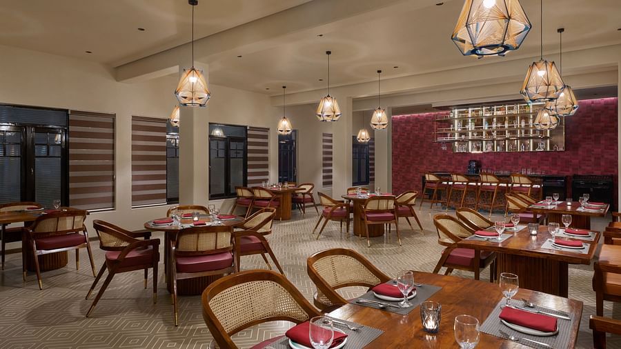Elegant indoor restaurant with warm lighting, wooden furniture, red cushioned chairs, and a bar area set against a textured maroon wall at Olive Restrobar, DGV Resort by Treat, Silvassa