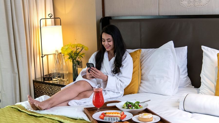 image of a woman scrolling through her phone sitting besides a tray served with a platter of breakfast - The Claridges New Delhi