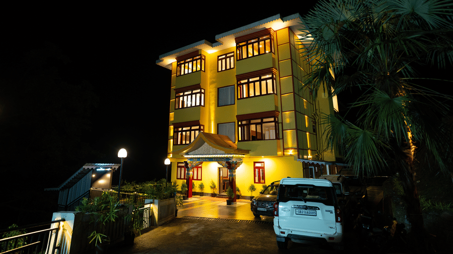 Night view of Voyage The Royal Retreat, a brightly lit yellow building with large windows, entrance canopy, parked cars, and surrounding greenery.