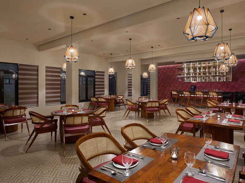 Elegant indoor restaurant with warm lighting, wooden furniture, red cushioned chairs, and a bar area set against a textured maroon wall at Olive Restrobar, DGV Resort by Treat, Silvassa