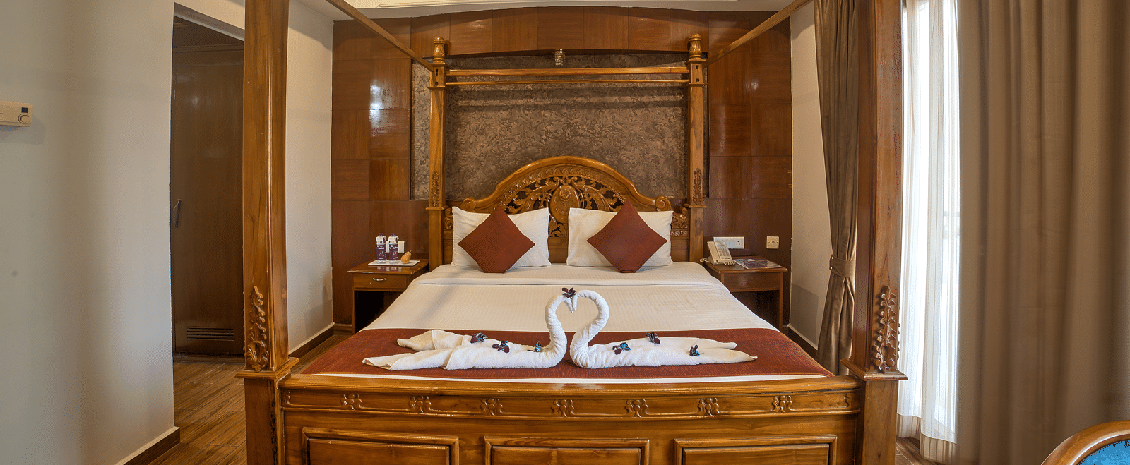 Classic wooden four-poster bed with decorative swan towel art in a warmly lit room at Ilara Hotels & Spa, OMR, Chennai