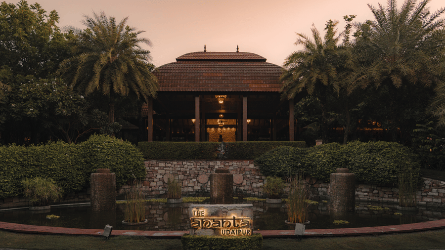 A building with a distinctive roof, possibly a resort or hotel, viewed across a body of water. Lush greenery and trees surround the building, creating a tranquil setting. The lighting suggests either dusk or dawn.